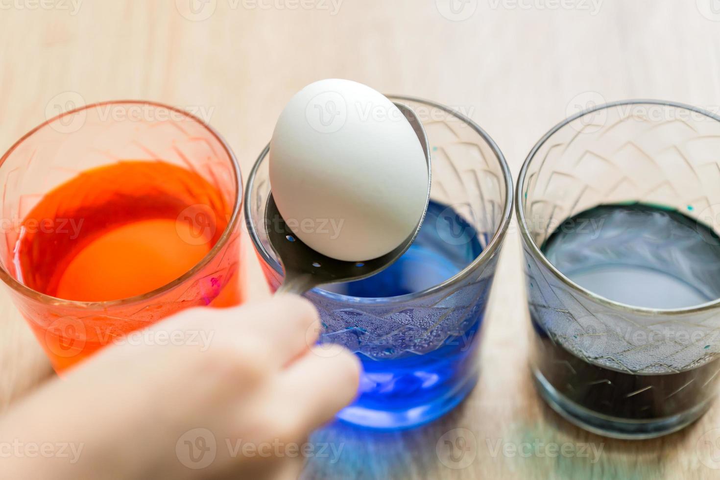 Transparent glass cups with colored water. The process of coloring eggs. A child hands put in a white egg with a spoon. photo