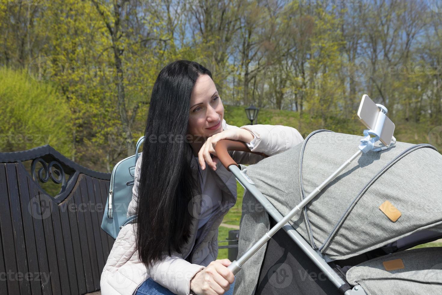 Young beautiful mother in dark hair, a woman with a cute baby in a pram makes a selfie from the phone. photo