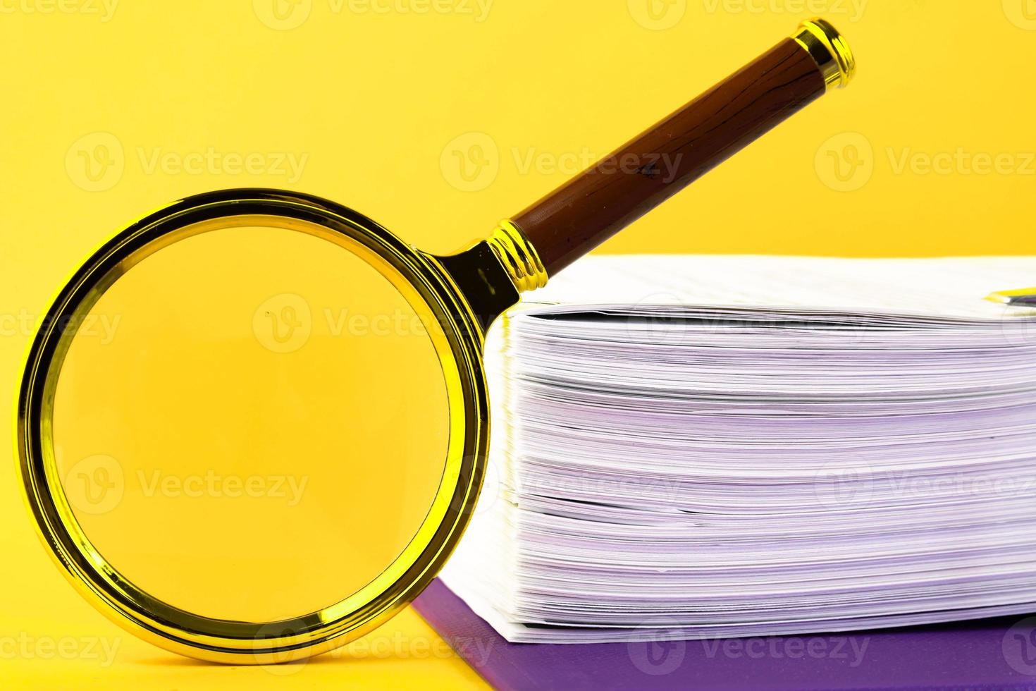 Close up of a pile of unfinished documents awaiting verification, a magnifying glass, on a yellow background. Business and education concept. Copy space photo