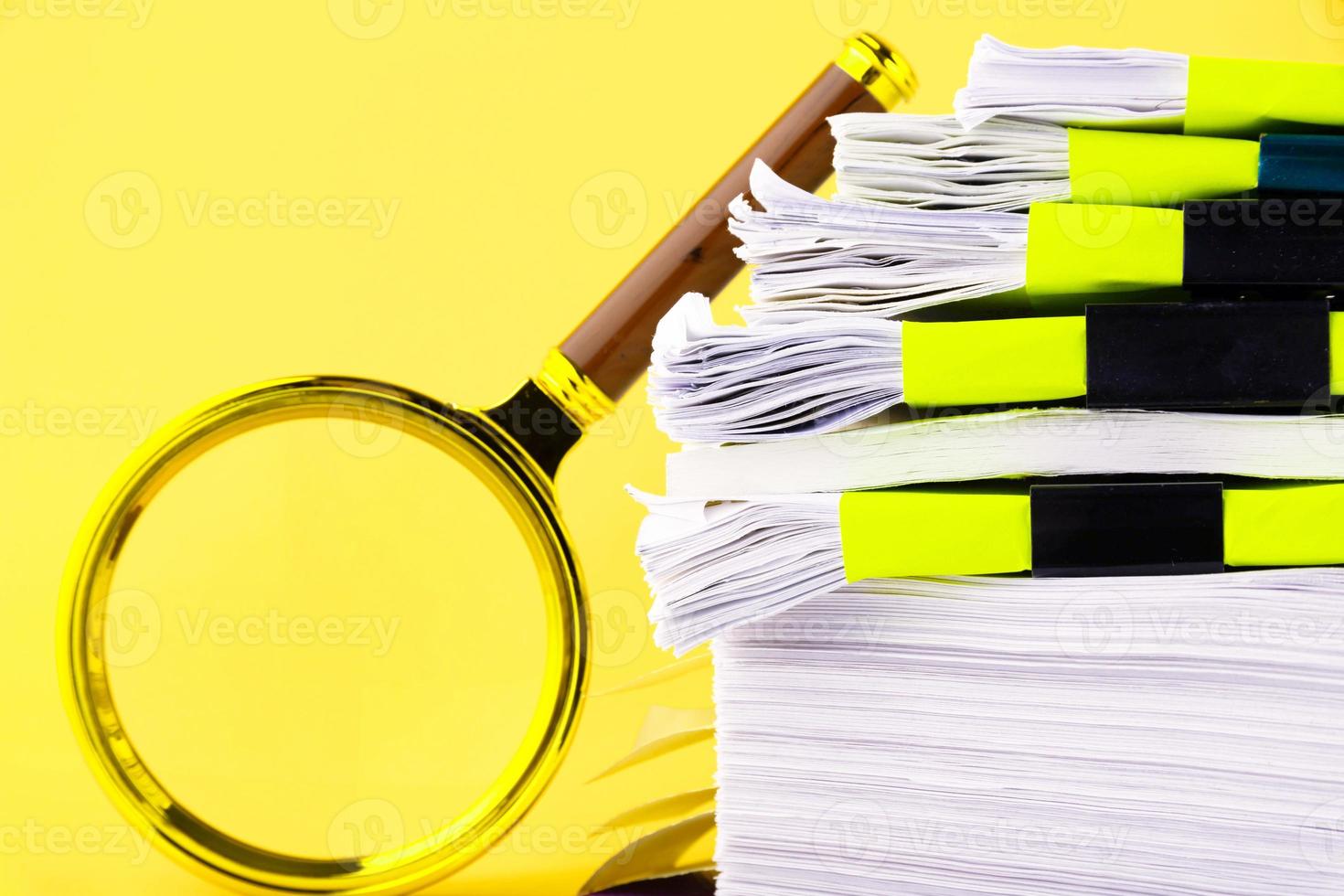 A large stack of unfinished documents awaiting verification, a magnifying glass, against a yellow background. Business and education concept. Copy space photo