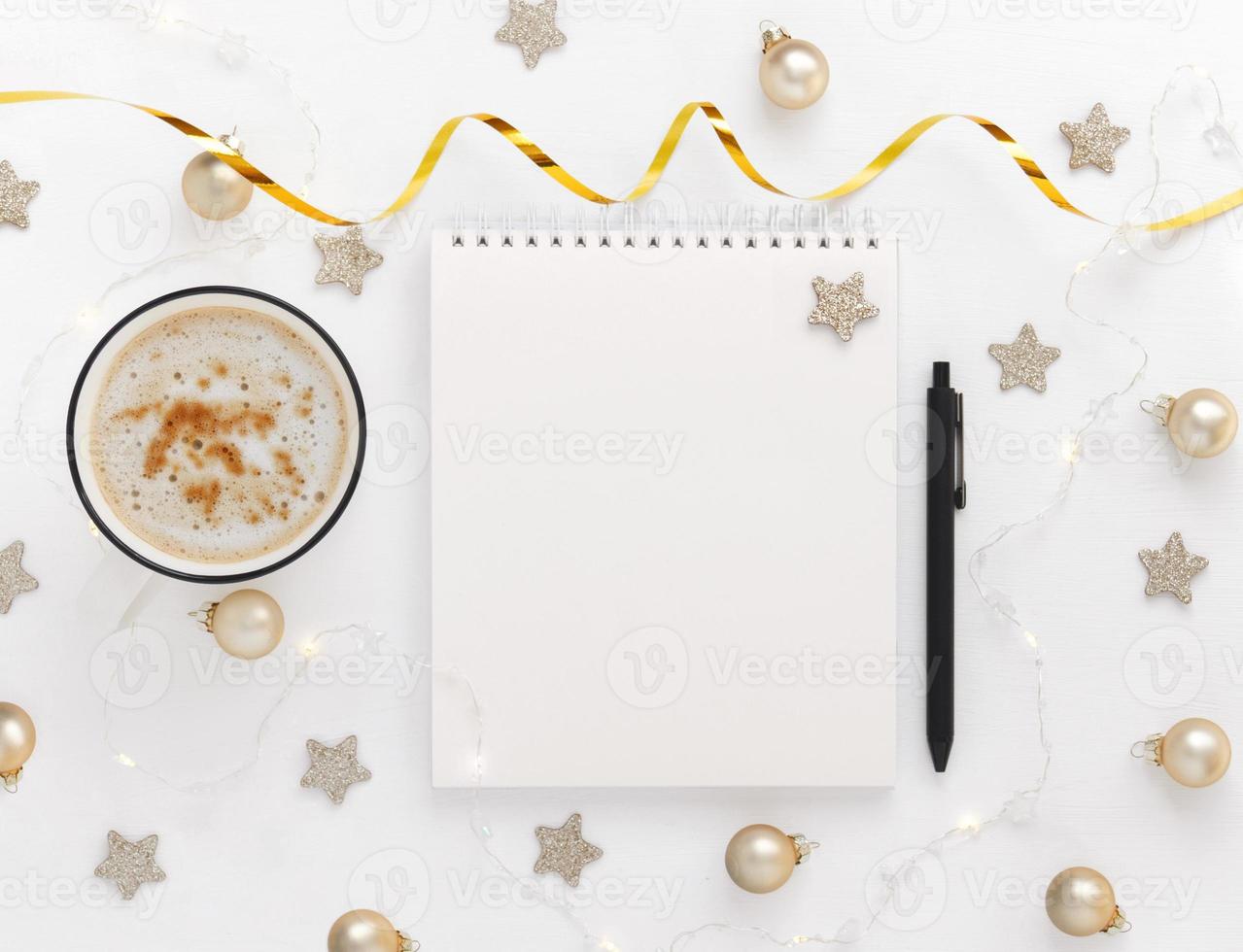 hoja blanca de cuaderno con bolígrafo sobre mesa blanca con taza de cacao o café, estrellas, bolas de navidad y guirnaldas festivas. planificación conceptual, lista de deseos para el nuevo año foto