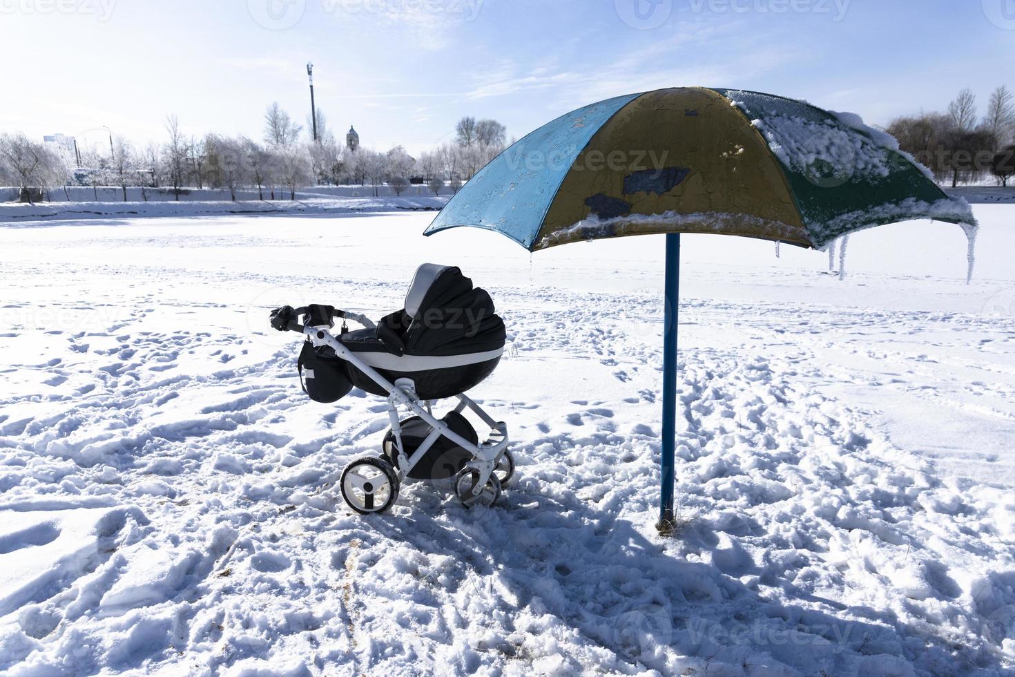 Baby carriage by the lake in winter. Beach under the snow and an umbrella from the sun. The concept of winter holidays with children. photo