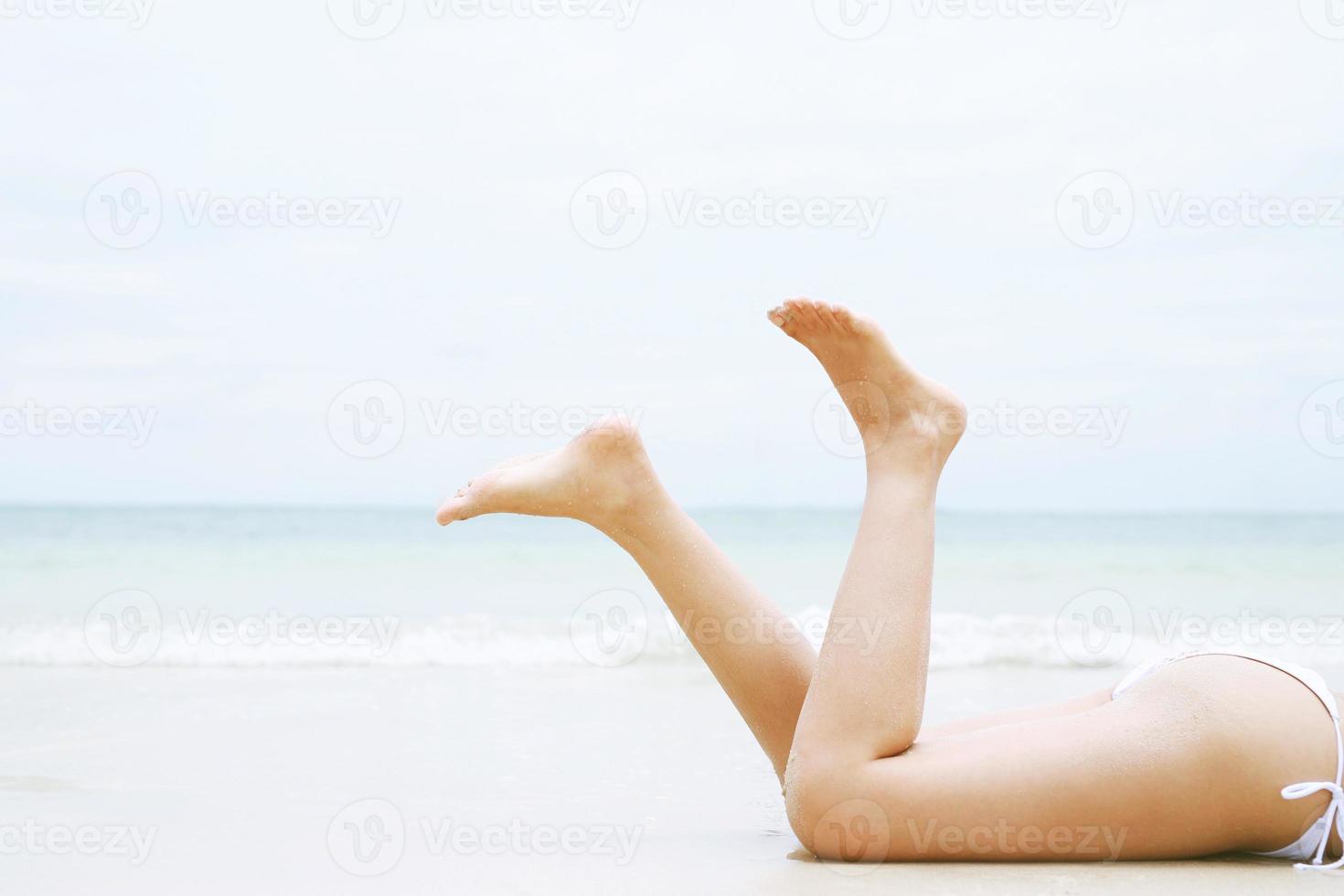 tanning suntanned young woman in bikini wearing straw hats lying on a tropical beach on white sand stretching up slender leg Blue sea in the background. Summer holiday fashion vacation travel concept. photo