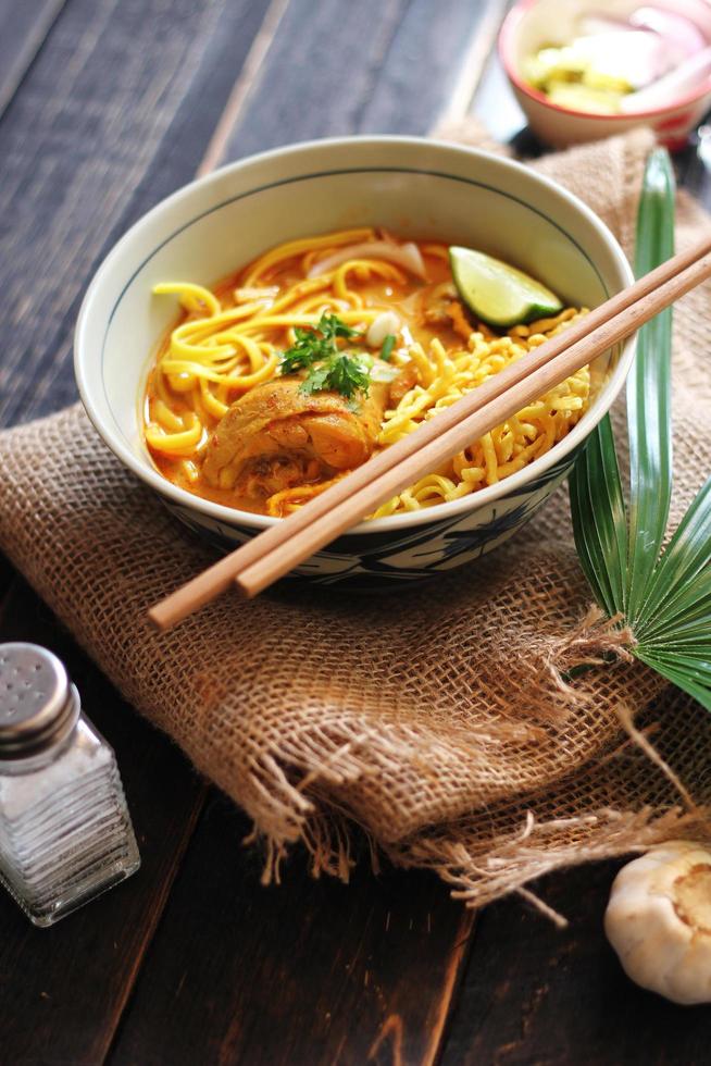 receta de khao soi, khao soi, khao soi kai, fideos tailandeses khao soi, curry de pollo con condimento servido en una mesa de madera negra con hojas decorativas, foto vertical.