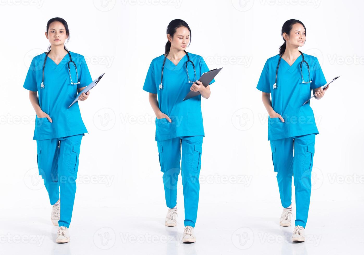 Mujer joven de 20 años de longitud completa, mezcla de raza, enfermera del hospital, caminando hacia adelante a la izquierda, paso a la derecha, con uniforme azul de estetoscopio. La doctora siente una sonrisa feliz sobre un fondo blanco aislado foto