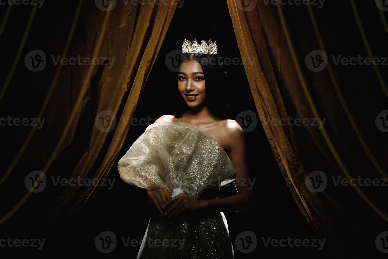 silueta de mujer de concurso de concurso de belleza miss con corona de diamantes brillantes con piel bronceada hermoso vestido de noche de maquillaje en el escenario con cortina de iluminación y fondo oscuro foto