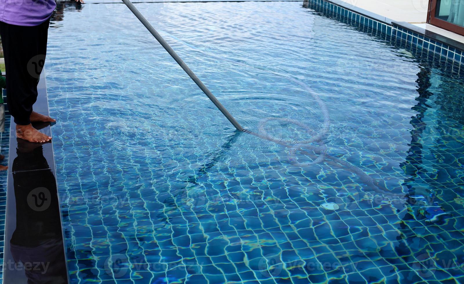 Woman Worker clean private swimming pool with vacuum blue tube cleaner every week in Summer, Salt type Pool along with Ocean as horizontal style with coconut trees photo