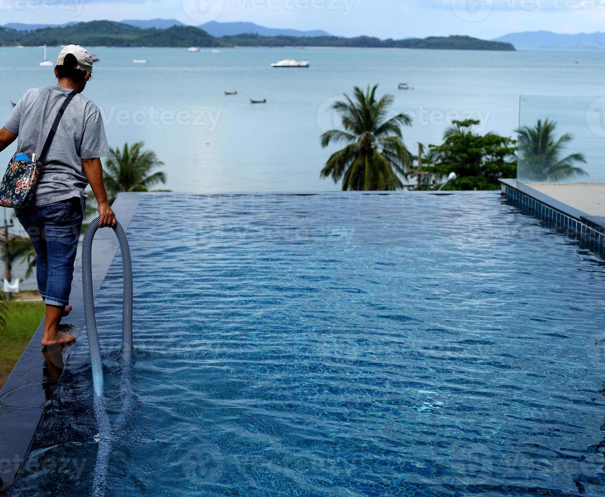 mujer trabajadora limpia piscina privada con limpiador de tubo azul al vacío cada semana en verano, piscina tipo sal junto con el océano como estilo horizontal con cocoteros foto