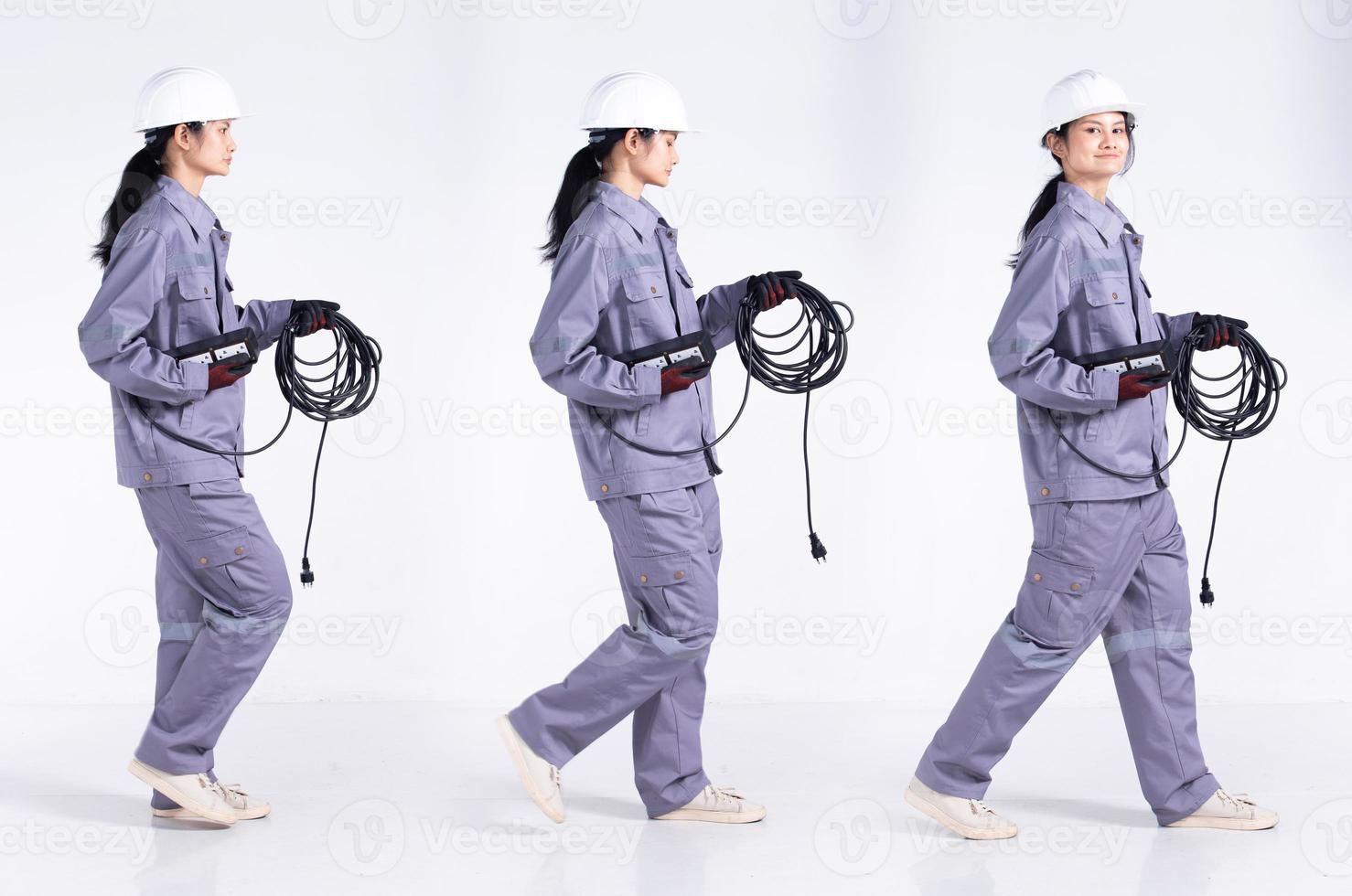 Full length 20s young Mix Race engineer electrician Woman, walking forward left right, wear gray uniform. Contractor female hold electric plug cable smile happy over white background isolated photo