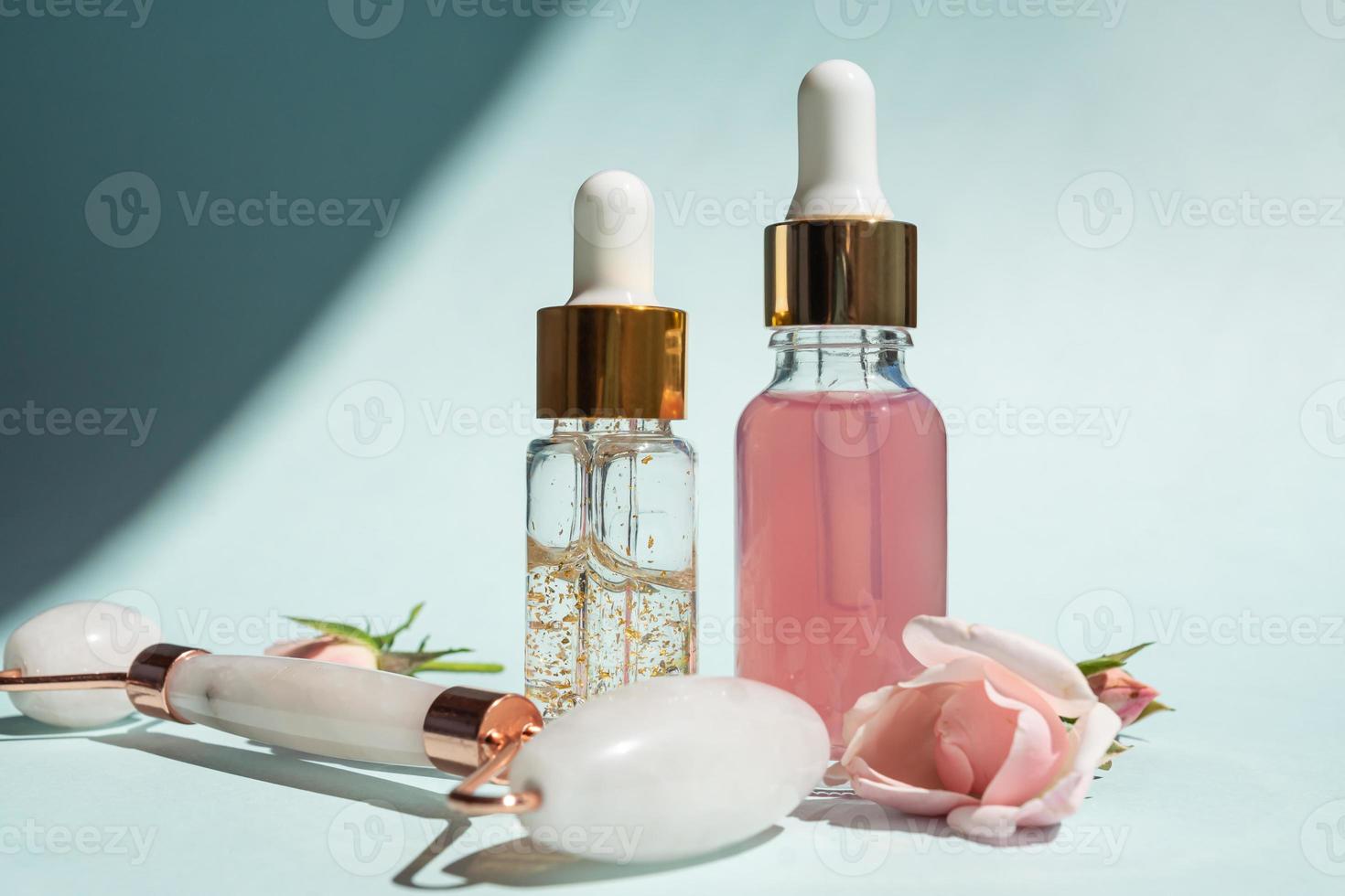 Massage roller for the face made of rose quartz with bottles of cosmetic oil or serum on a blue background. The concept of skin care at home. 24k gold serum and rose water for face treatment. photo