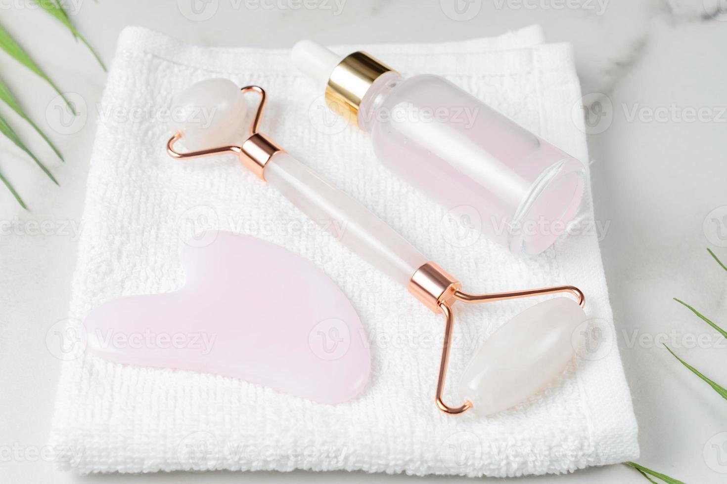 Pink quartz Gua sha scraper and roller face massager and bottle of cosmetic serum for the face on white towel. photo