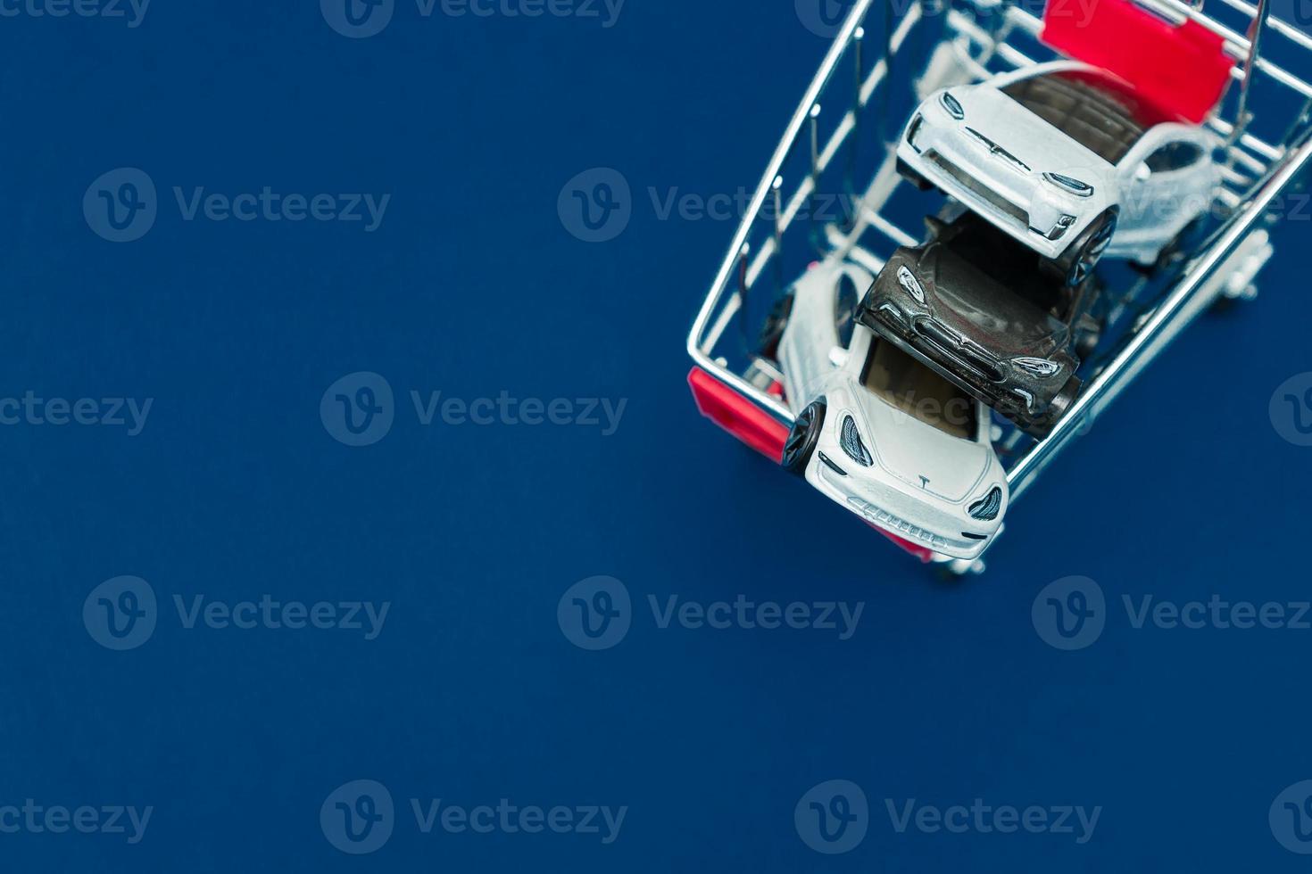 luxury toy cars Tesla models in small shopping cart on classic blue background. Concept of buying new luxuary car photo
