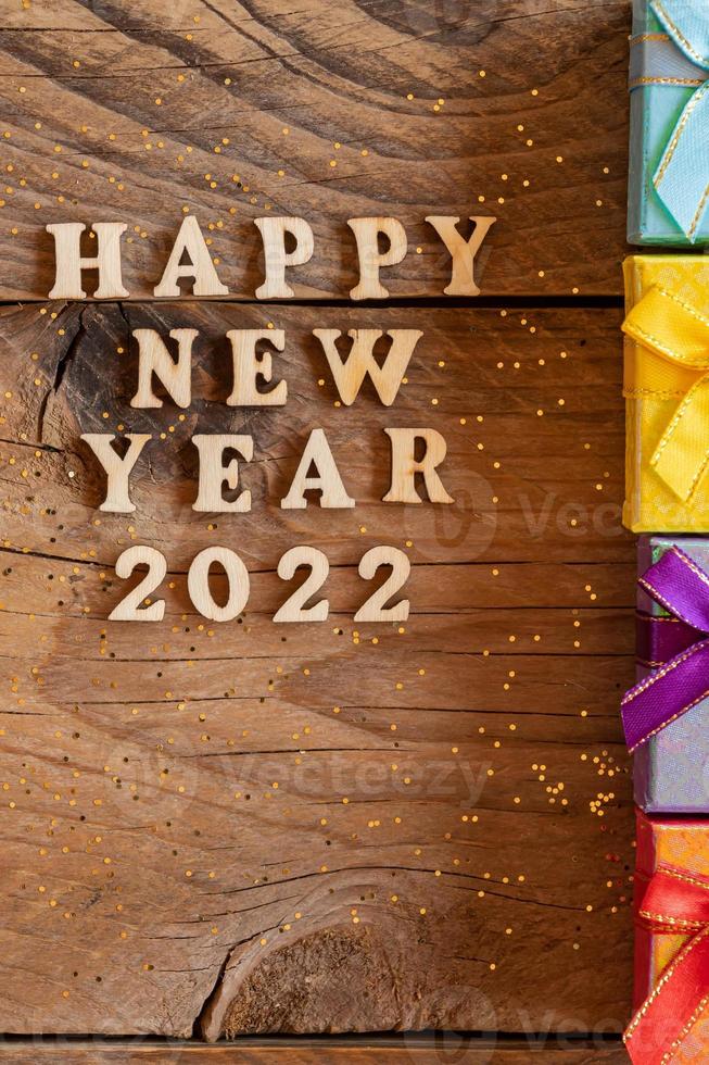 Happy New Year 2022. Quote made from wooden letters and numbers 2022 on wooden background with multicolored smal gift boxes. Creative concept for new year greeting card photo