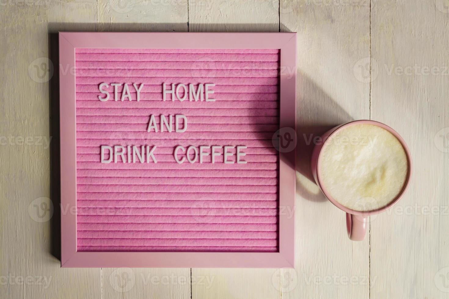 top view of Pink cup with cappuccino and qoute Stay home and drink coffee. Self isolation and quarantine campaign to protect yourself during coronavirus pandemia. photo