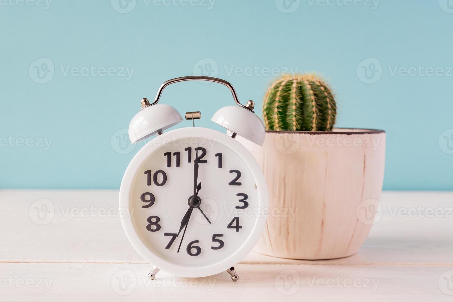 White alarm clock standing on a wooden shelf on green backdrop. Cactus in a pot on the background photo