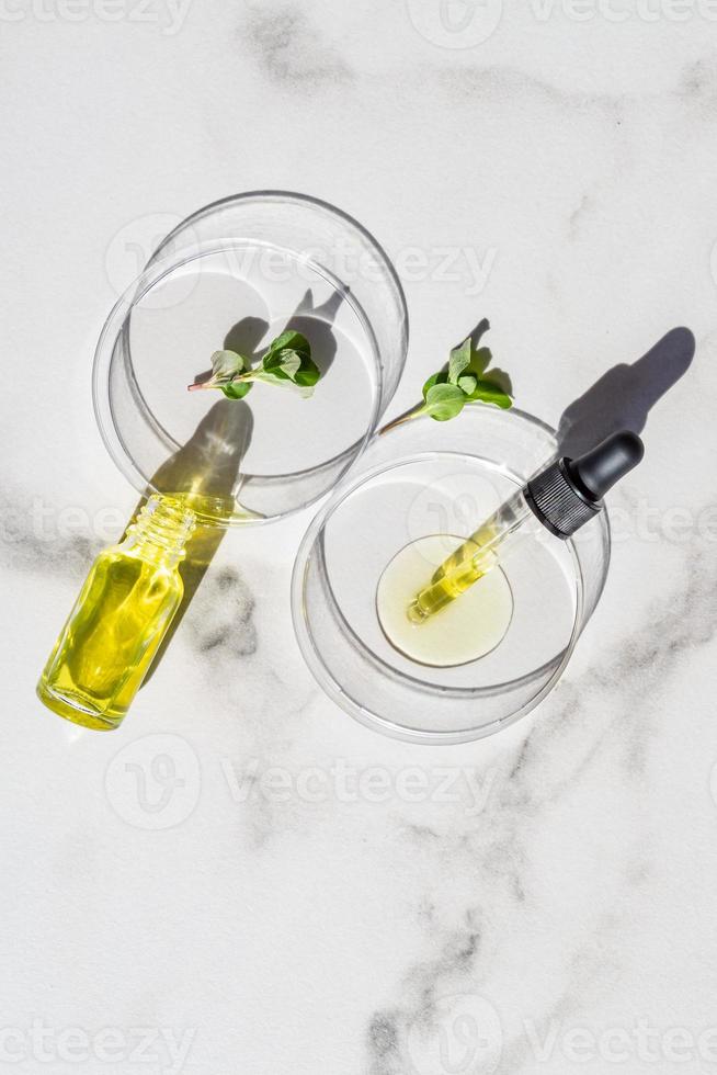 suero de pipeta cosmética en una placa de petri con hojas de menta verde sobre fondo de mármol blanco. concepto de cosmetología y productos farmacéuticos. peeling ácido, aceite facial, colágeno foto