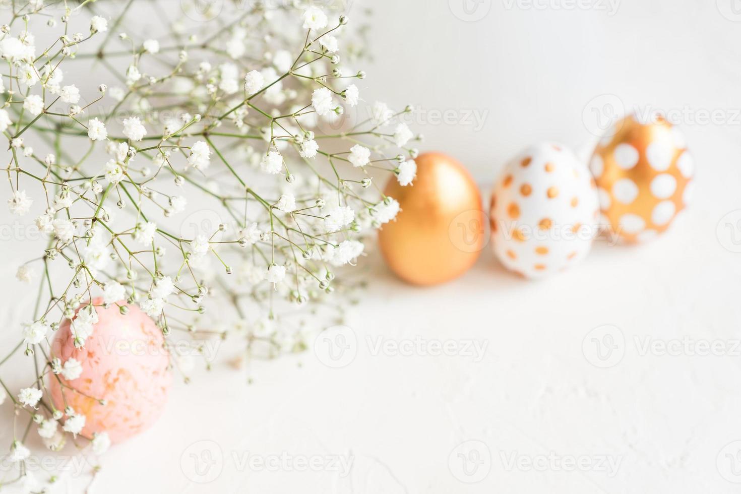 Close up tender blooming gypsophila flowers on background of rose, white and golden decorated easter eggs on white concrete background. Copy space for text photo