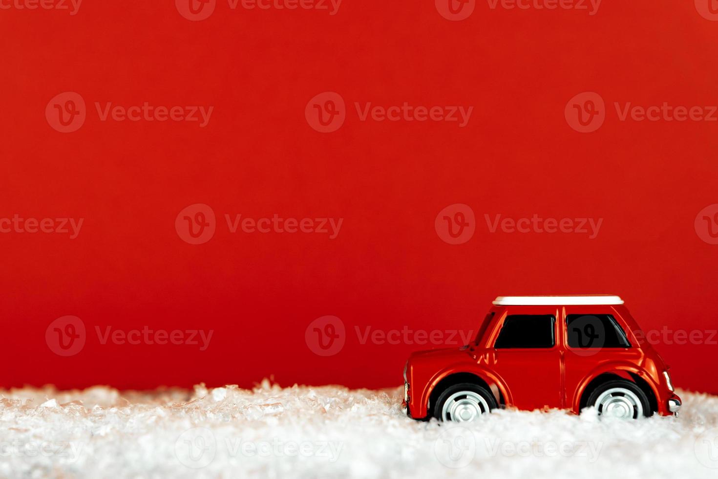 Small red toy car drives on a snowy road on red background. Festive christmas greeting card with copy space. photo