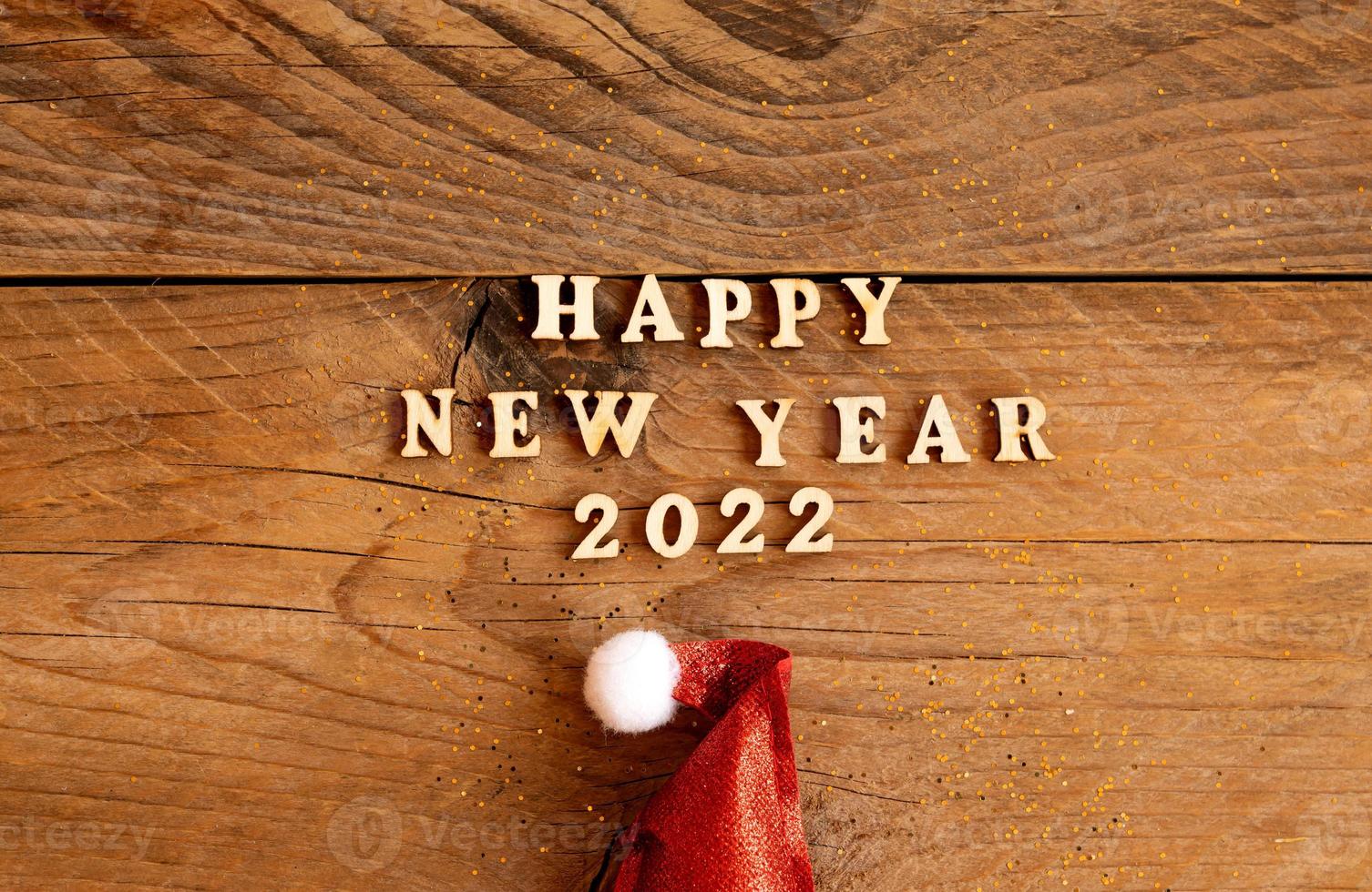 Happy new year 2022 wooden letters quote with red sparkling santa hat on wooden table with glitter. Festive greeting card for a new year celebration photo