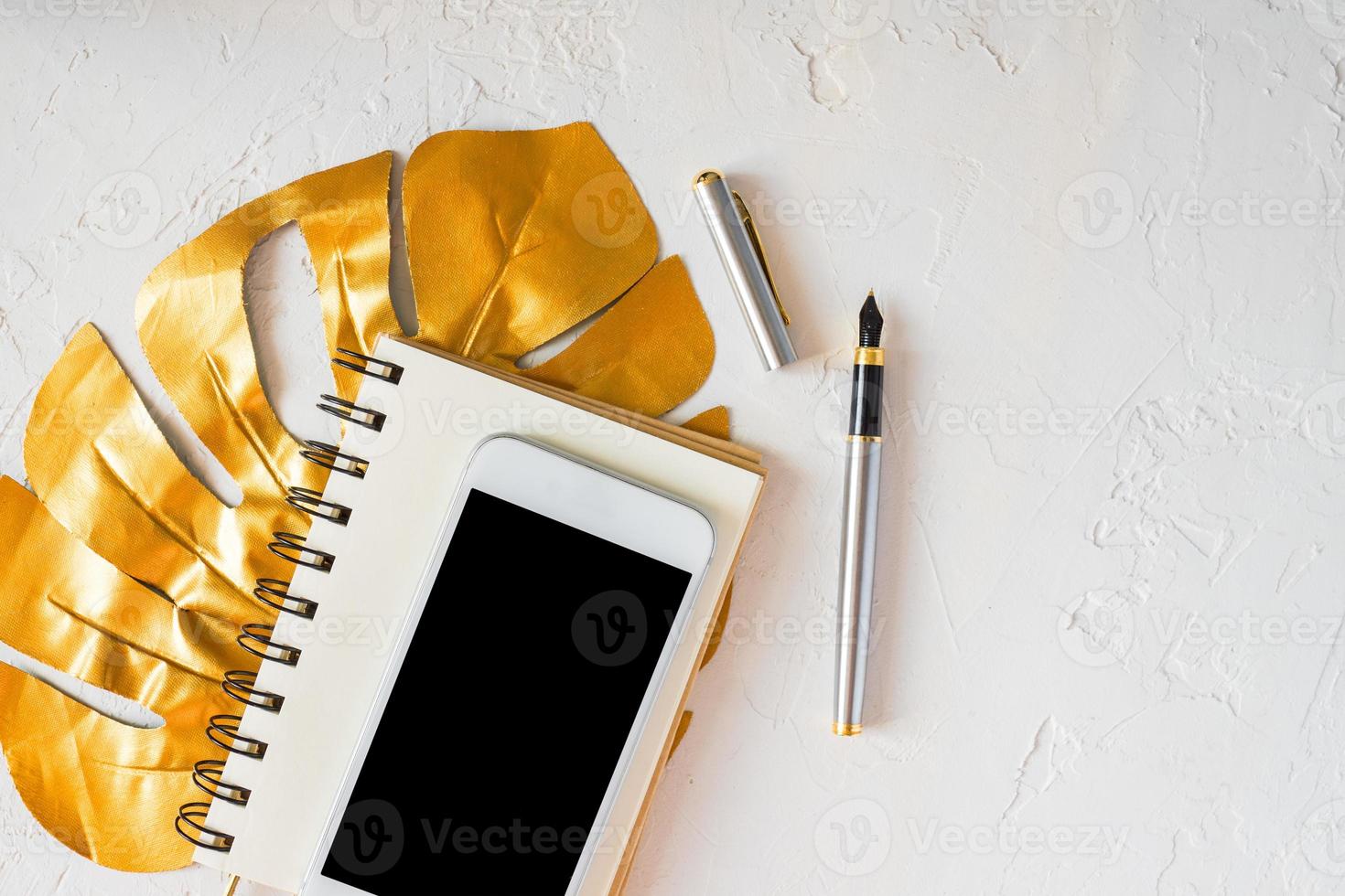 escritorio de mujer. hoja de monstera tropical dorada, bloc de notas en blanco, teléfono inteligente blanco y bolígrafo sobre fondo de hormigón blanco. foto