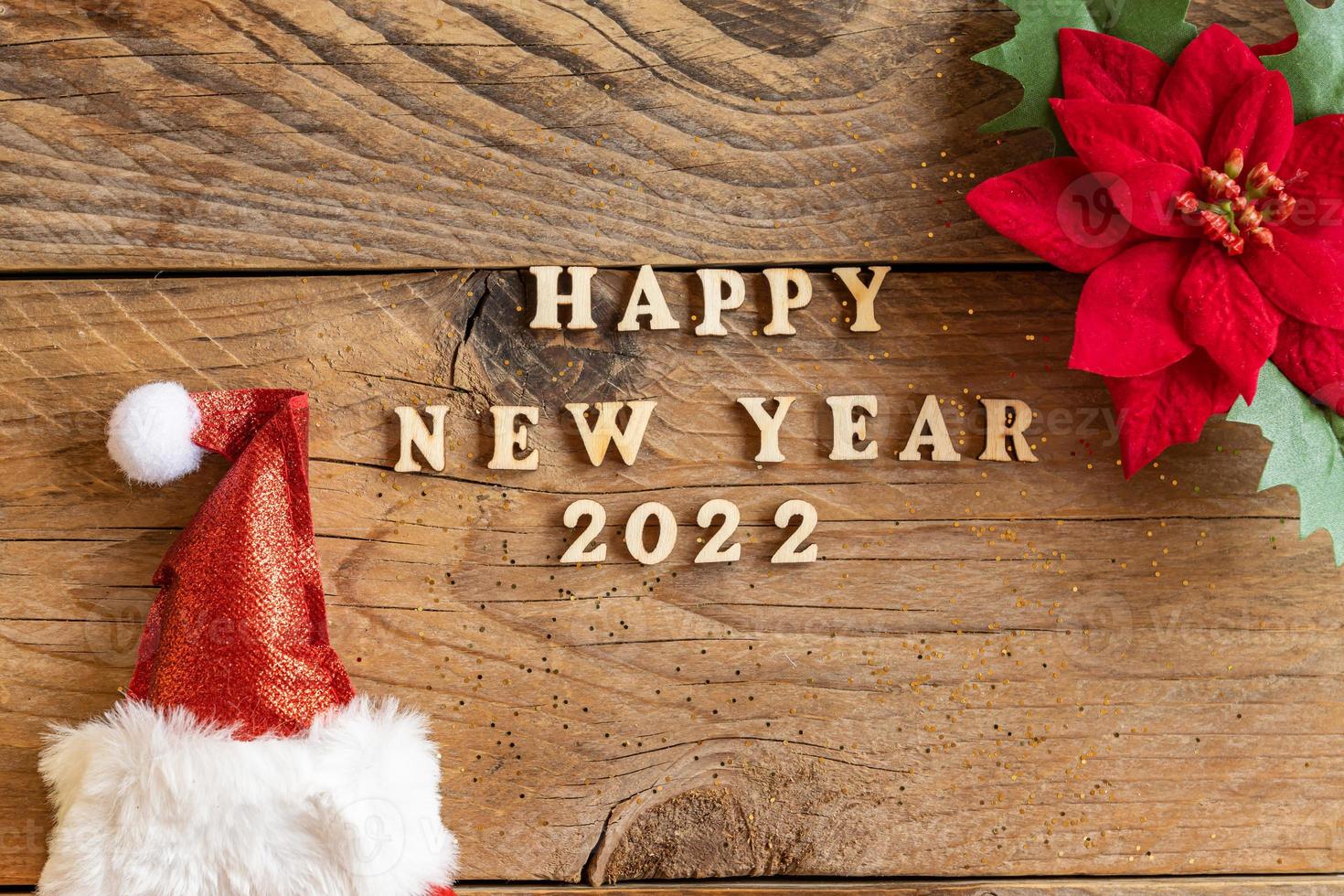 Happy New Year 2022. Quote made from wooden letters and numbers 2022 on wooden background with santa hat and poisentia flower. Creative concept for new year greeting card photo