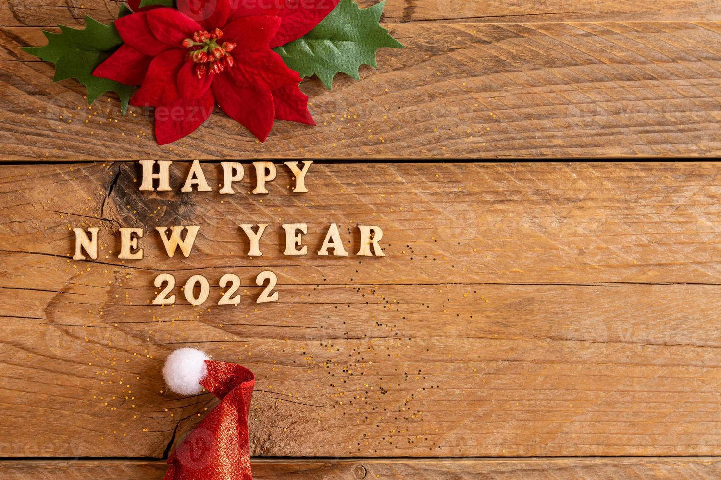 Happy New Year 2022. Quote made from wooden letters and numbers 2022 on wooden background with santa hat and poisentia flower. Creative concept for new year greeting card with copy space photo