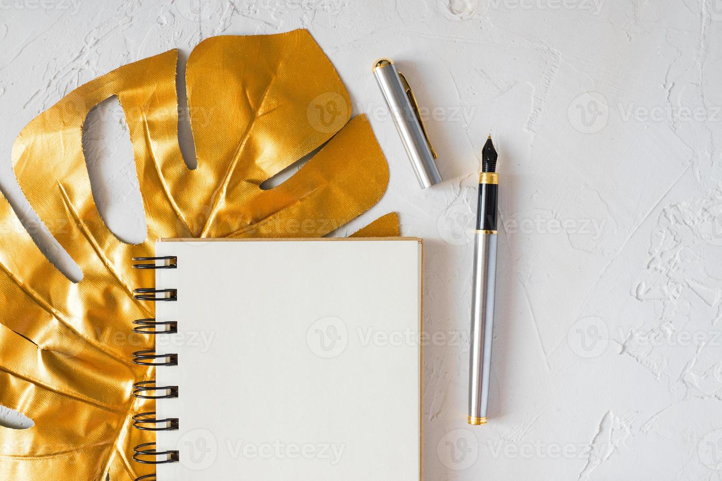 Stylish home office desk with paper notepad, golden pen and tropical monstera leaf. Flat lay, top view, copy space. Feminine workspace concept. photo