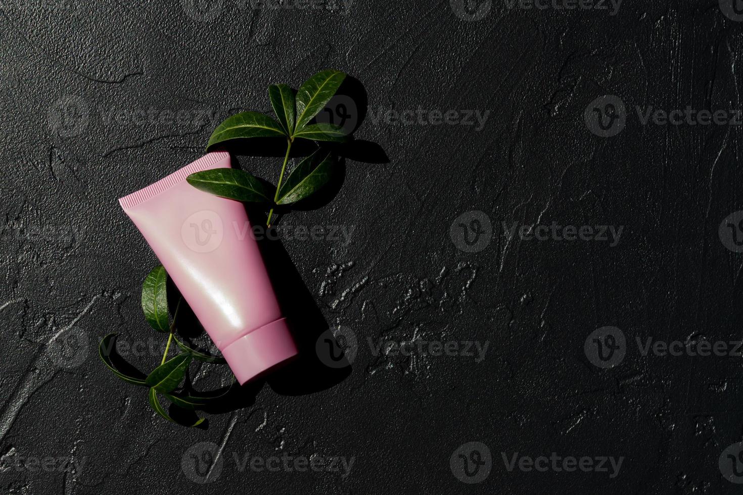 tubo cosmético rosa con crema facial, loción corporal o limpiador sobre un fondo oscuro con hojas verdes frescas. concepto de cuidado de la piel sensible. copia espacio, maqueta foto