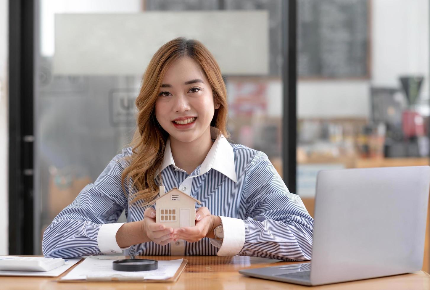Miniature house in the hands of an Asian woman real estate agent home loan working at the office. Looking at the camera. photo