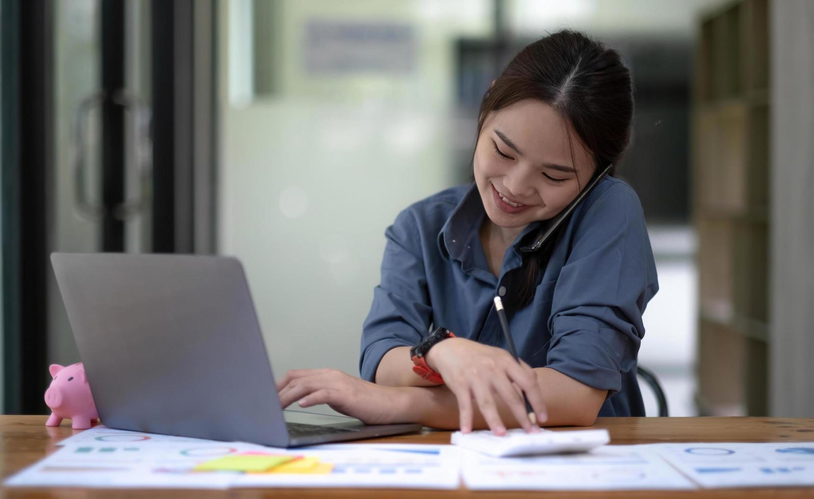 Portrait of Asian young female talking on the phone with a business customer while using a calculator recording data on her work computer. photo