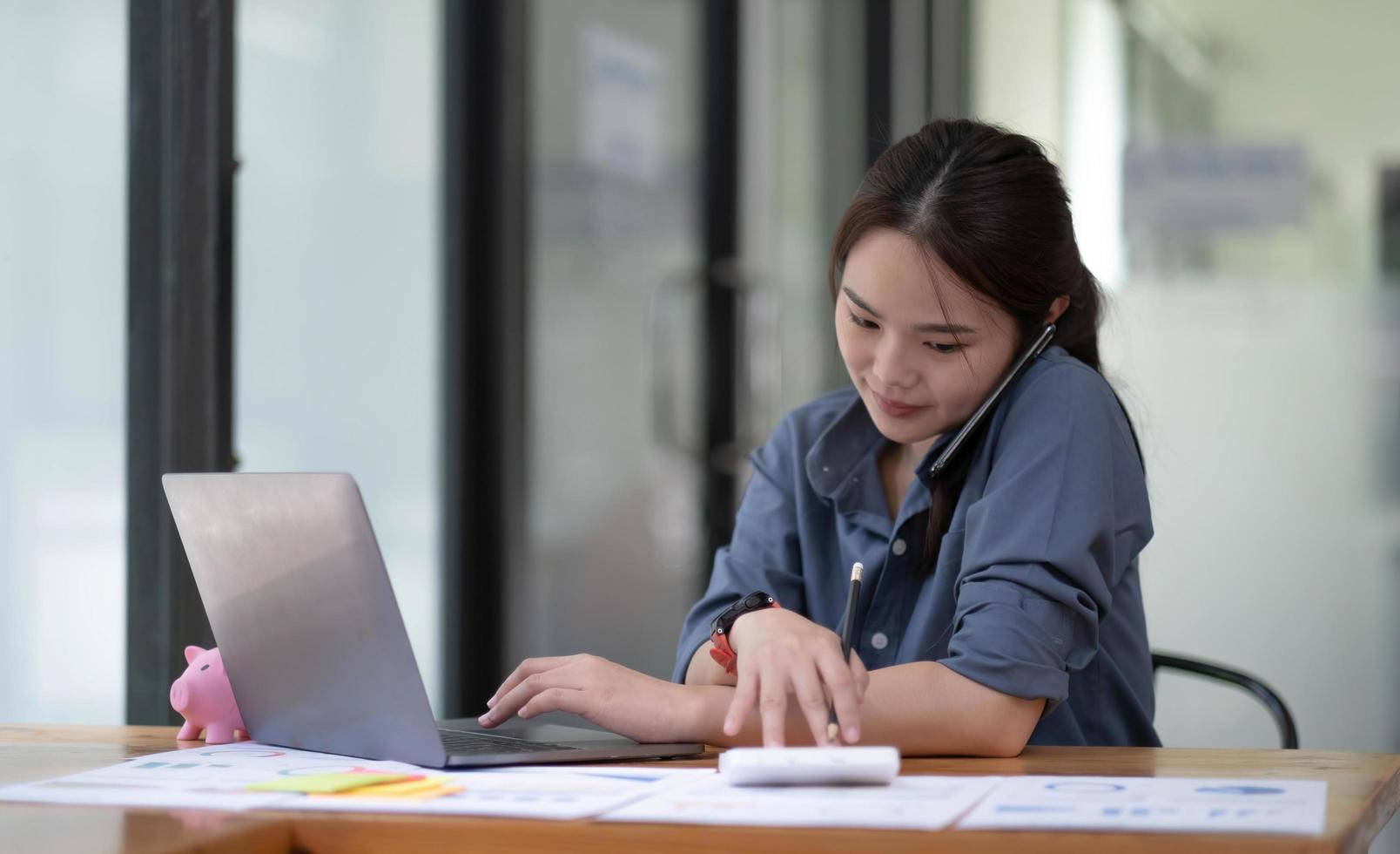 Asian female manager talking on the phone with a business customer while using a calculator recording data on her work computer. photo