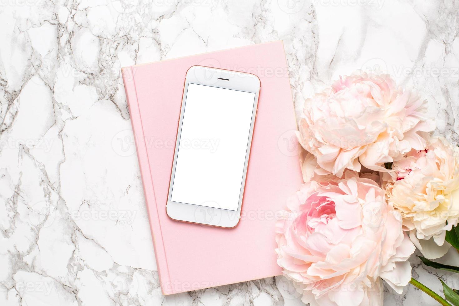 Mobile phone with a white and pink notebook and piony flowers on a marble background photo