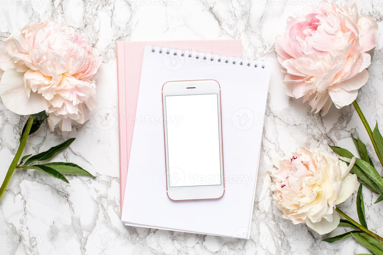 Mobile phone with a white and pink notebook and piony flowers on a marble background photo