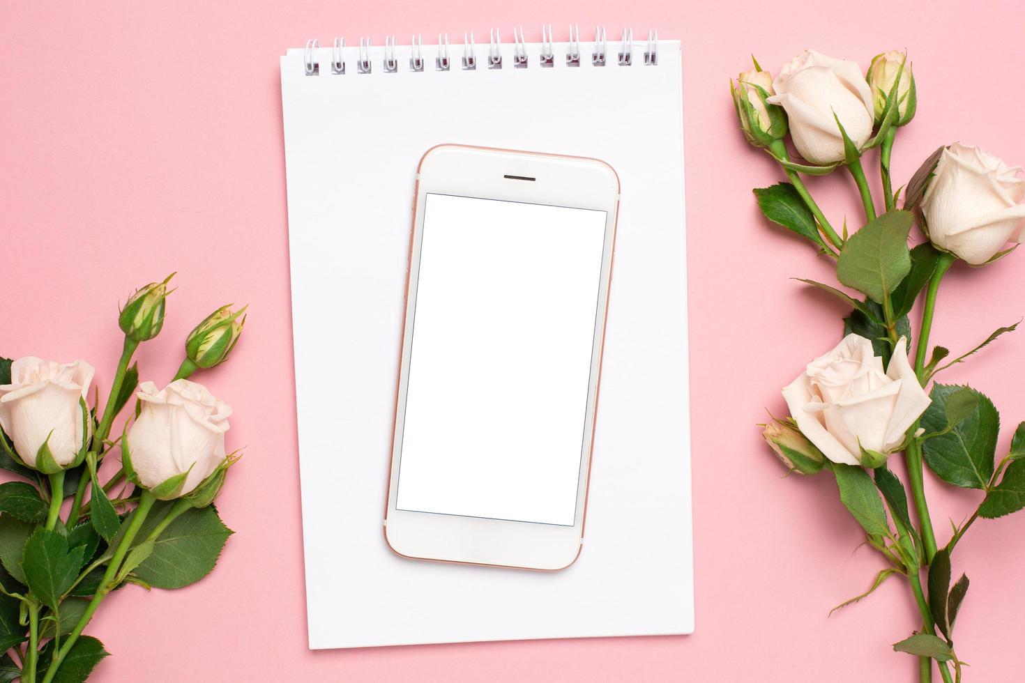Mobile phone with a white notebook and roses flowers on pink background photo