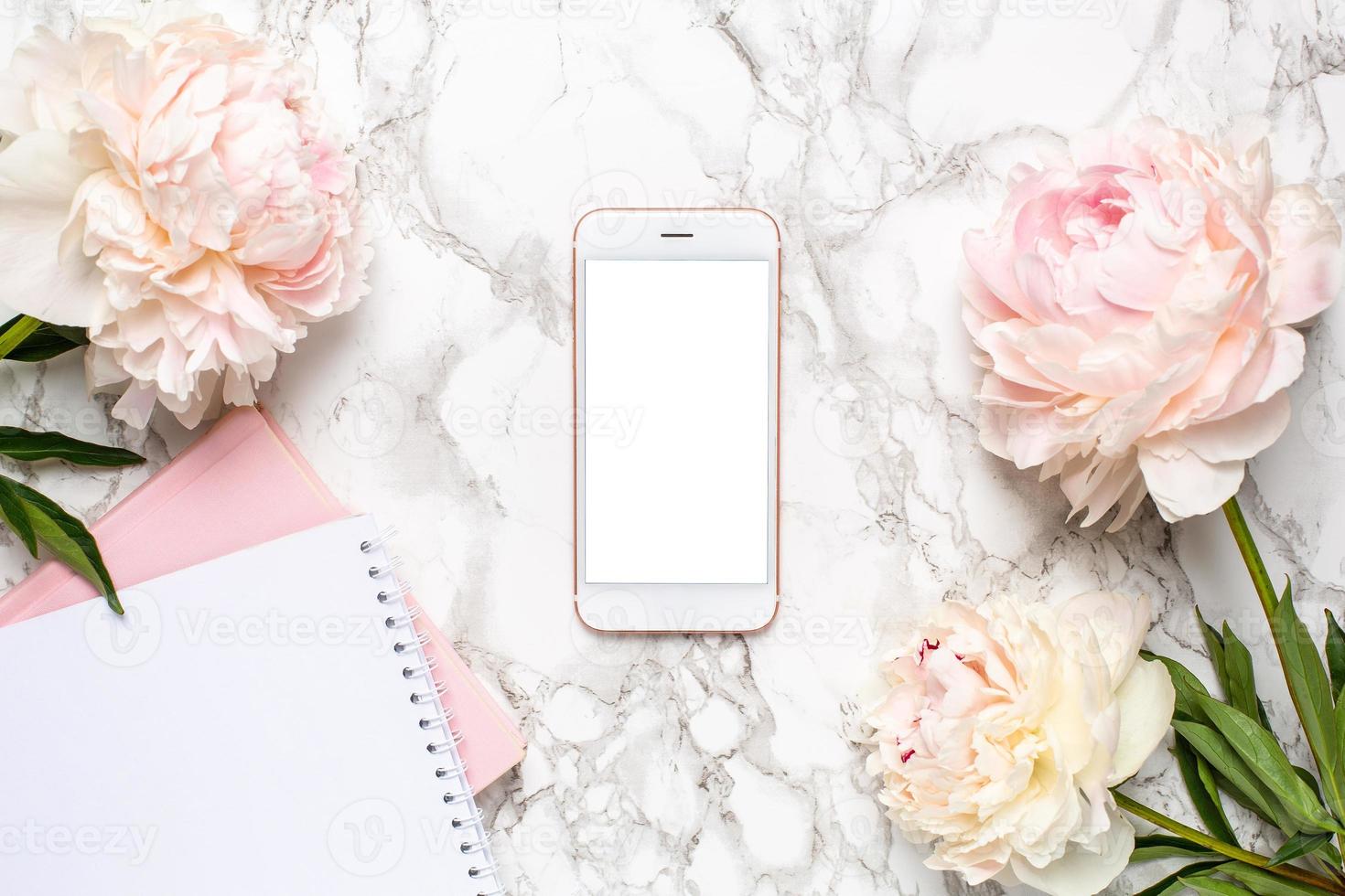 Mobile phone with a white and pink notebook and piony flowers on a marble background photo