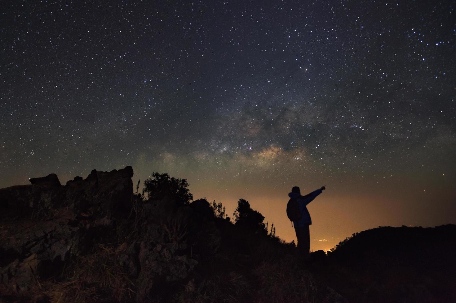 un hombre está de pie junto a la galaxia de la vía láctea apuntando a una estrella brillante en doi luang chiang dao con signos de punto superior en idioma tailandés. fotografía de larga exposición con grano foto