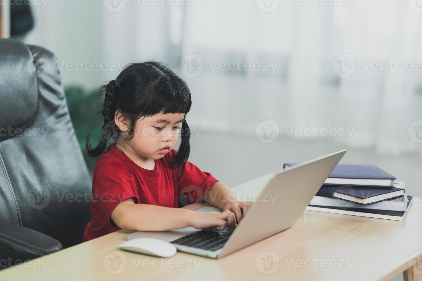 una niña asiática que usa una camiseta roja usa una computadora portátil y estudia en línea en un escritorio de madera en la sala de estar en casa. educación aprendiendo en línea desde el concepto de hogar. foto