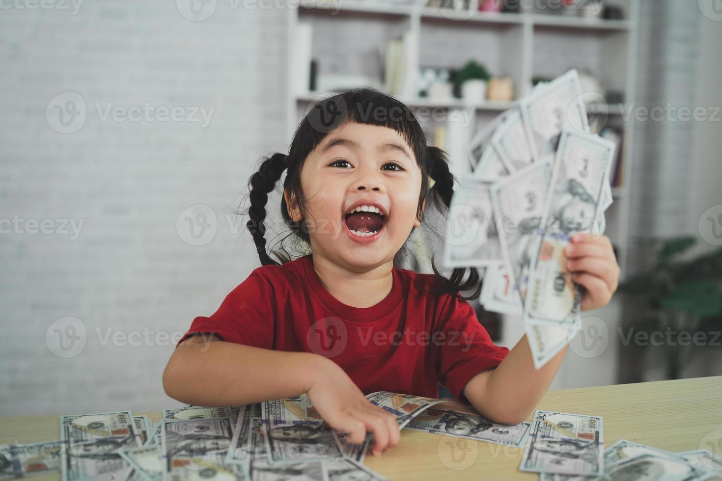 niña asiática con una camiseta roja sosteniendo un billete de un dólar en una mesa de madera en el salón de casa. concepto de riqueza de inversión de ahorro. foto