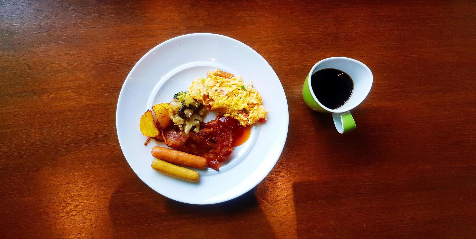Top view of breakfast with omelet, bacon, many sausage, fried vegetable and tomato or chili sauce with green cup of black coffee. Delicious meal in American style with hot drinking on wooden table. photo