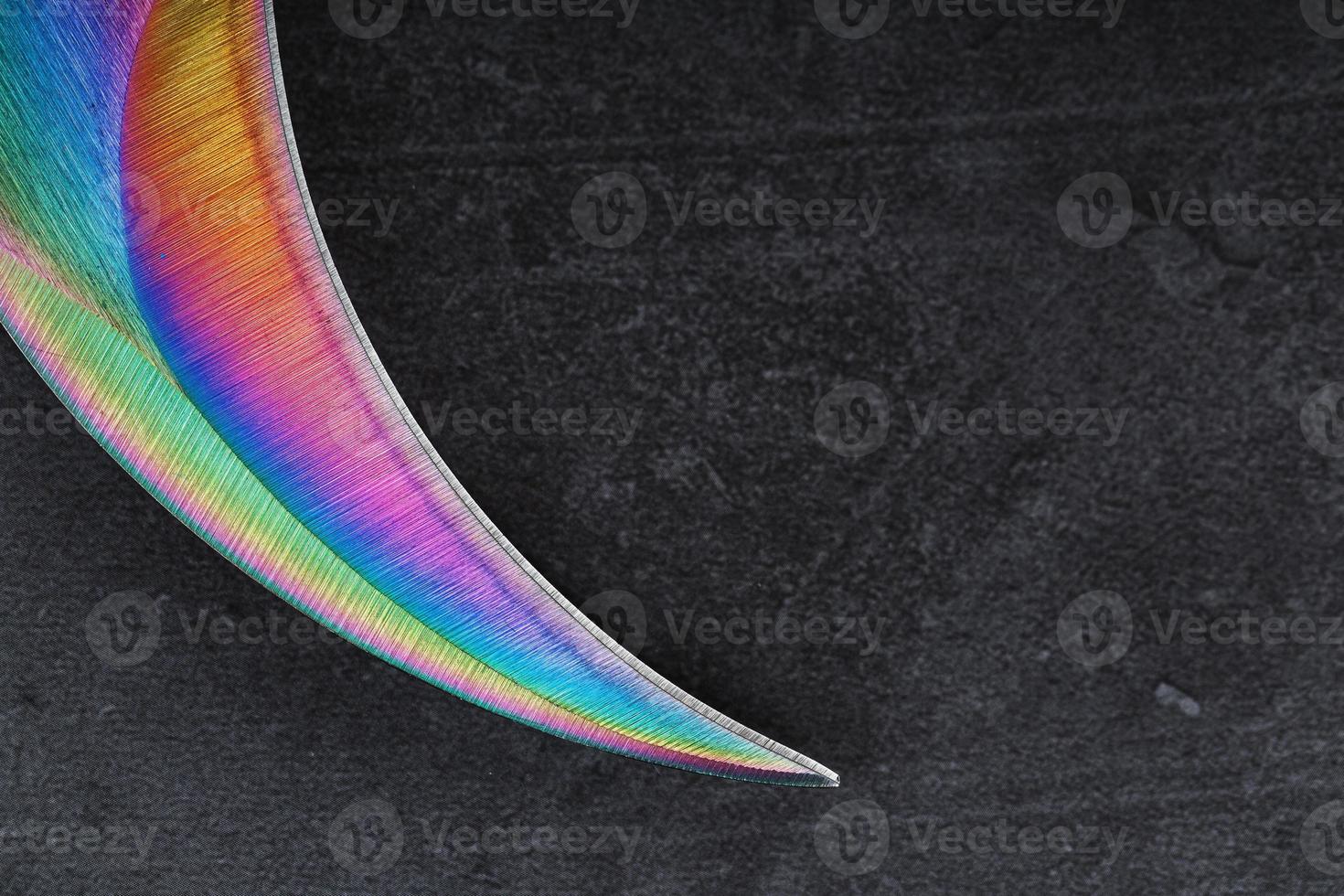 The curved sharp blade of the Kerambit Dagger is a gradient rainbow color on a dark background. photo