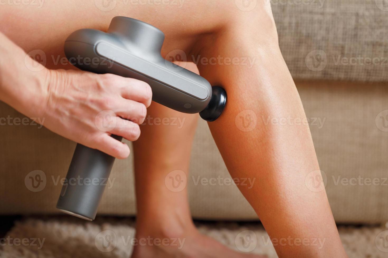 una mujer masajea sus piernas con una pistola automática en casa. foto