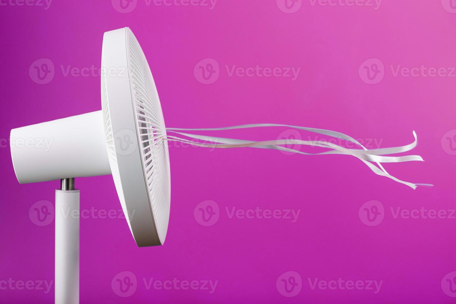 The electric fan is white with pink ribbons fluttering in the wind on a pink background. photo