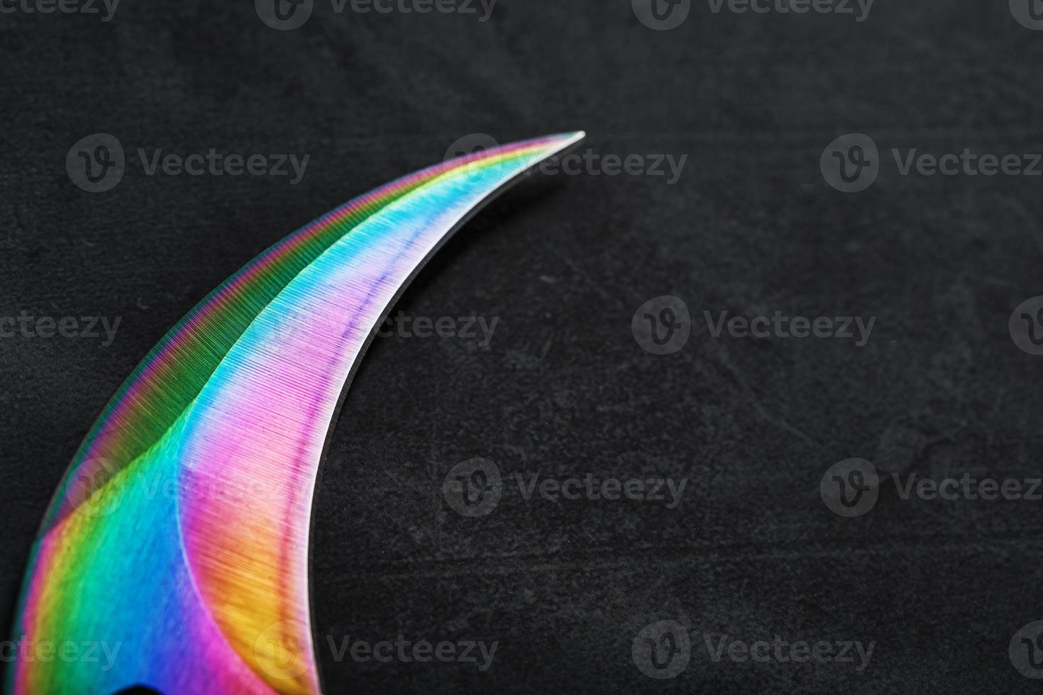 The curved sharp blade of the Kerambit Dagger is a gradient rainbow color on a dark background. photo