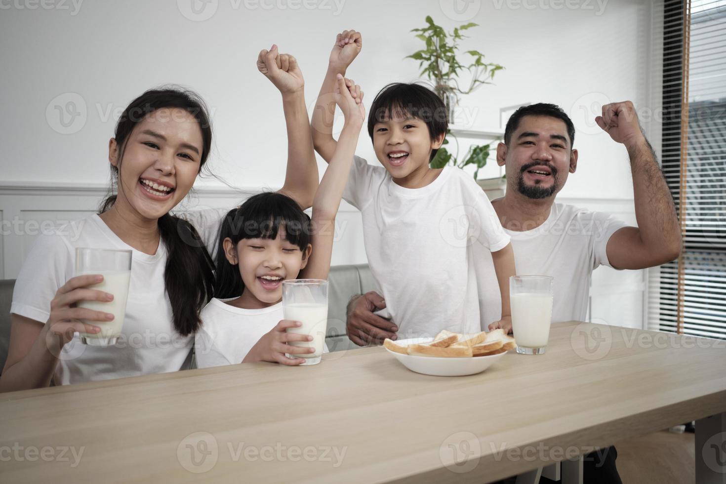 una familia tailandesa asiática saludable, niños pequeños y padres jóvenes beben leche blanca fresca en un vaso y pan de alegría juntos en una mesa de comedor en la mañana, bienestar nutrición hogar desayuno comida estilo de vida. foto