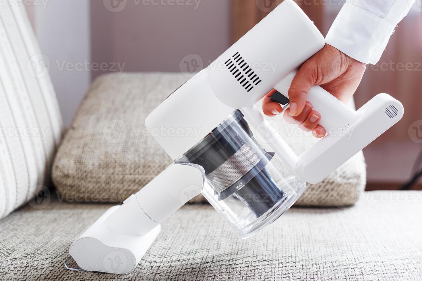 A white cordless vacuum cleaner in a woman's hand cleans the sofa in the house in close-up. photo