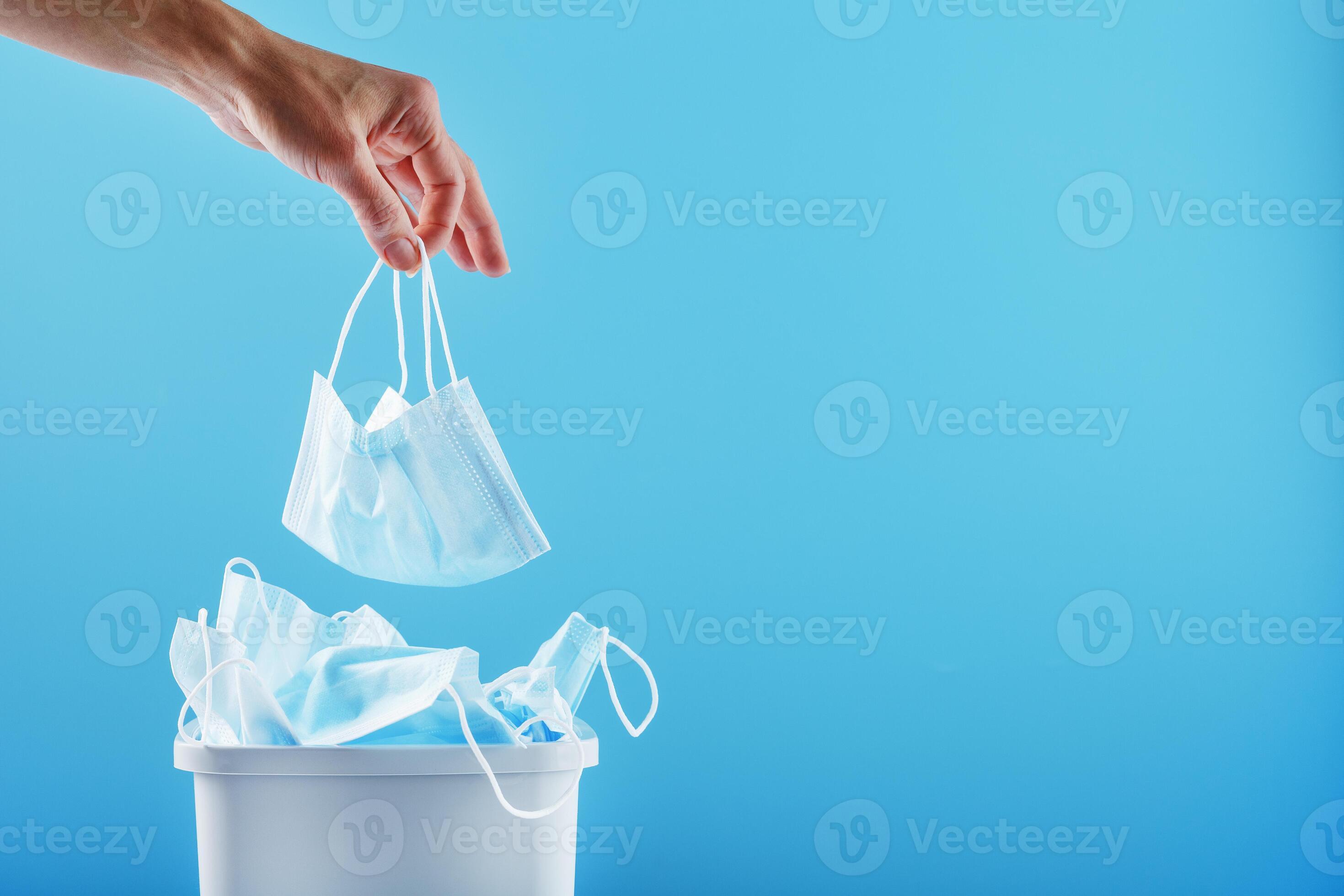 A hand throws a used protective mask into a trash can filled with masks from a virus on a blue
