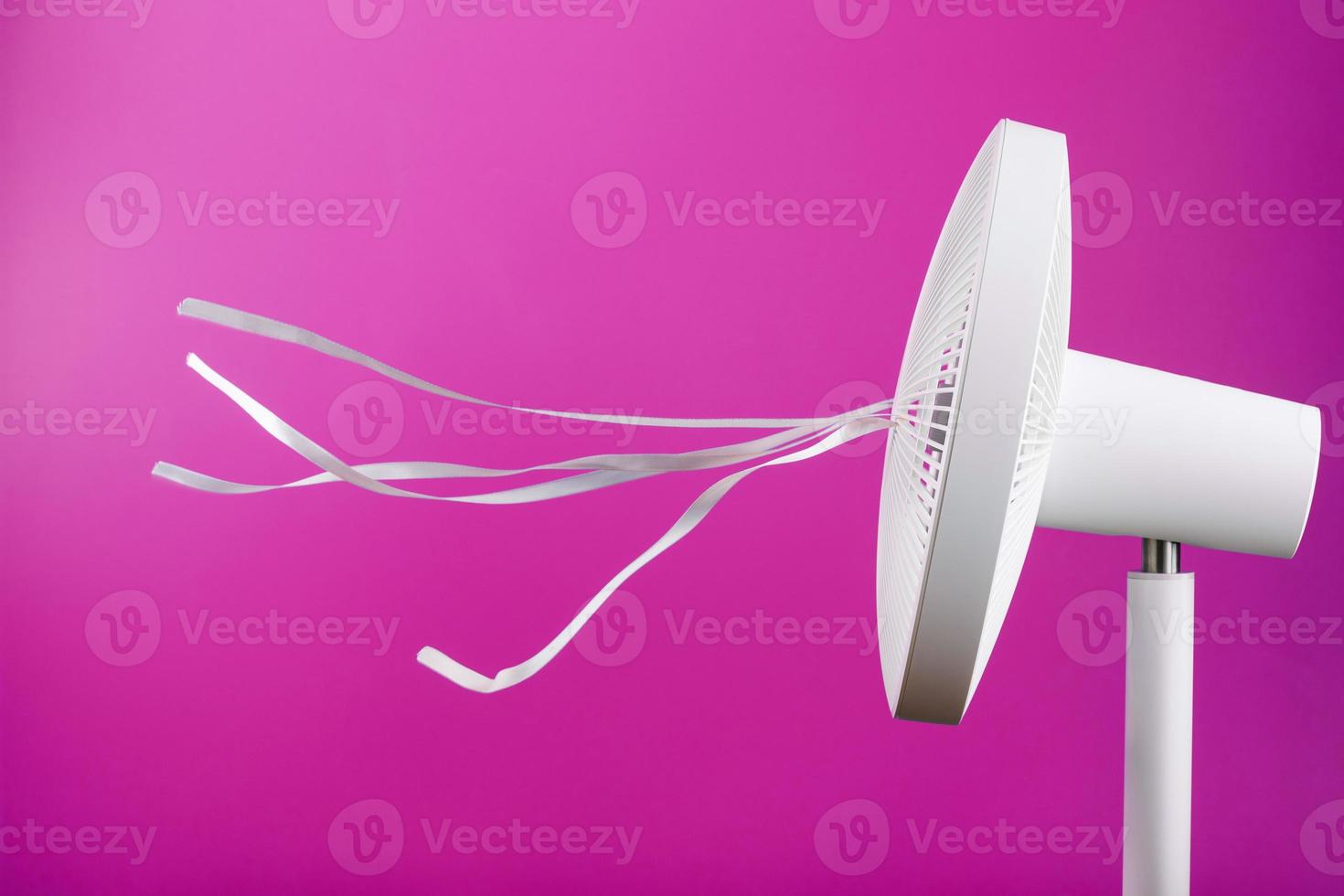 The electric fan is white with pink ribbons fluttering in the wind on a pink background. photo