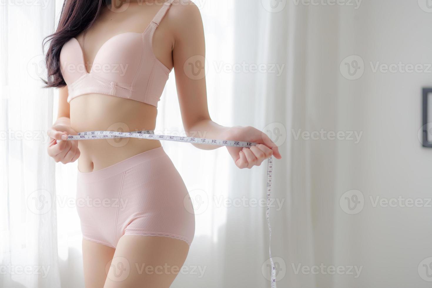 hermosa mujer asiática joven cuerpo sexy cuerpo delgado que mide el abdomen para controlar la pérdida de peso en la habitación, el vientre de la chica de belleza tiene celulitis con cinta métrica para el concepto de dieta, salud y bienestar. foto