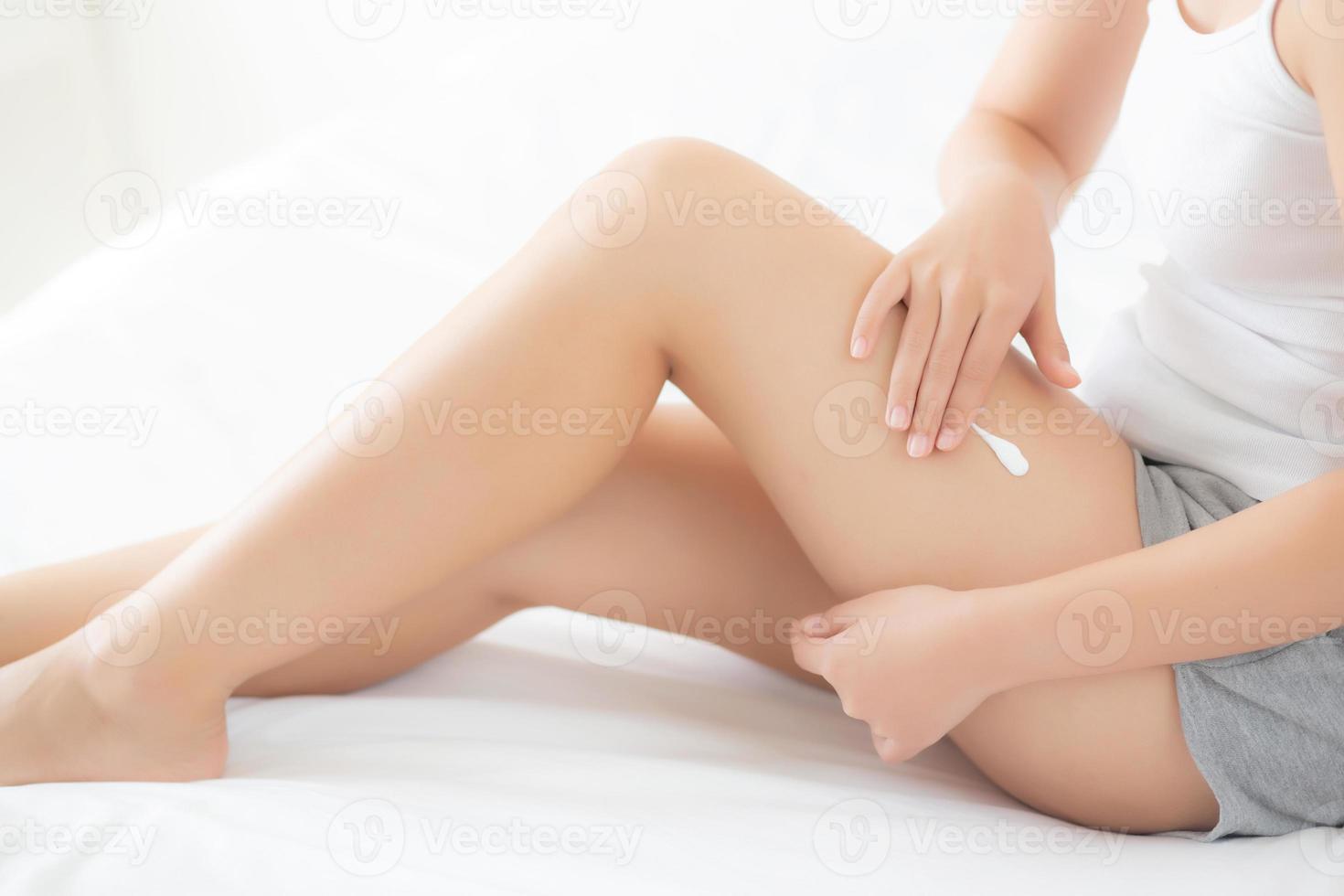 Beautiful young asian woman sitting on a bed stroking legs with soft smooth skin in the bedroom, girl applying touch body cream and lotion with treatment care, healthy and wellness concept. photo