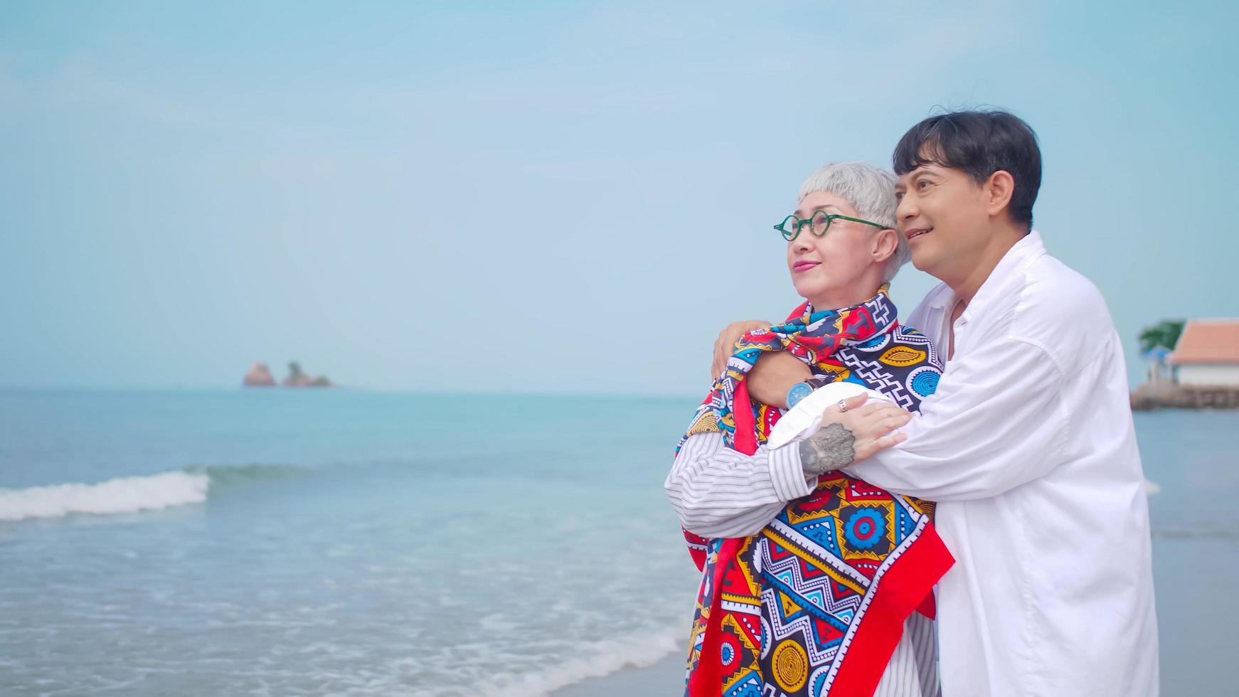 Asian retired couple relaxing by the sea in summer. A senior man taking care to his partner while standing on the beach. photo